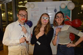Two young women and a man are Standing in front of a poster which Shows the Eifeltower and the Television Tower. The are dressed with French Accessoires like beret, moustache and scarves.